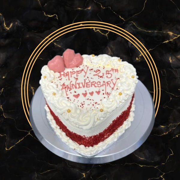 Heart-shaped red velvet anniversary cake with white cream frosting, golden pearls, and pink chocolate hearts