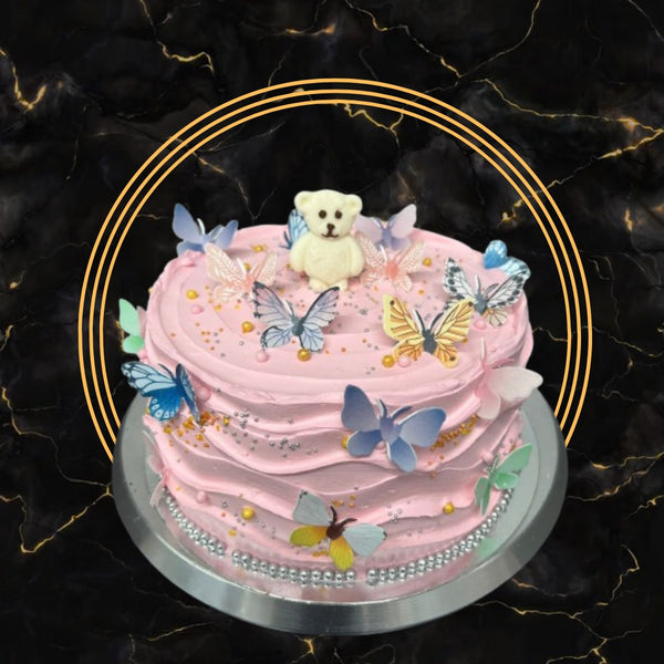 Pink cake decorated with edible butterflies, silver pearls, and a teddy bear topper