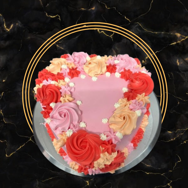 Pink heart-shaped cake decorated with red, peach, and pink buttercream flowers on a silver cake board